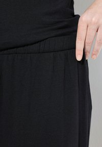 Zwarte kleding gemaakt van zachte stof, met een elastische tailleband en plooien voor extra textuur. Detail van een hand die op de tailleband rust.
