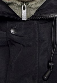 Schwarze Jacke mit einem Metallreißverschluss, Druckknopfverschluss und verstellbarem schwarzen Kordelzug mit einem Kunststoffstopper in der Nähe des Kragens.