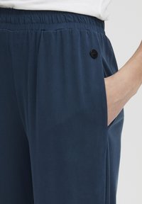 Marineblå bukser med en glat overflade, elastisk talje og sidelommer. Har en cirkulær sort knapdetalje nær lommen.