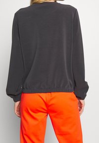 Felpa leggera grigia scura con maniche lunghe, vita elastica e polsini, abbinata a pantaloni arancioni vivaci. Texture liscia.