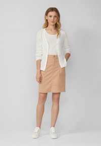 Witte geribbelde cardigan over een witte tanktop, gecombineerd met een beige aansluitende rok met een voorzak. Model draagt witte sneakers.