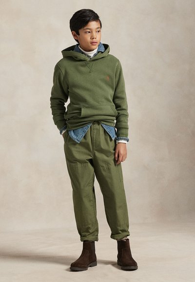 Olivno zeleni kapučino z žepom spredaj, prelit čez denim srajco. Olivno zeleni hlače in rjavi gležnjarji dopolnjujejo outfit.