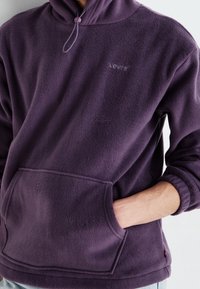 Persona che indossa una felpa con cappuccio viola Levi's in pile con tasca frontale, mano sinistra nella tasca, vista fino a metà lunghezza.