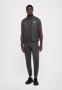 Gilet matelassé gris sur une chemise à manches courtes gris foncé, jogging gris assorti et baskets noires avec une semelle texturée. Logo EA7 visible.