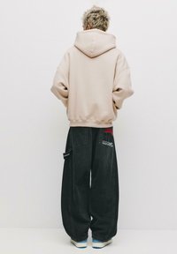 Hoodie oversize beige chiaro con un grande cappuccio, abbinato a jeans scuri e larghi con toppe e ricami. Sneakers bianche completano il look.