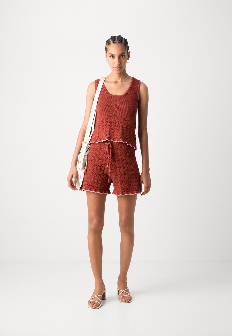 Rotes, strukturiertes Stricktanktop mit gewelltem Saum, kombiniert mit passenden Shorts. Das Model trägt helle Sandalen und hält eine kleine weiße Tasche.