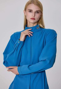 Chemisier bleu à col montant avec de longues manches bouffantes, présentant un détail de bouton au col et un tissu lisse et texturé.