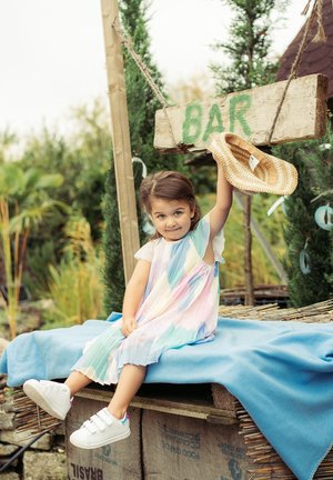 Bunt gestreiftes Kleid in Pastellfarben, sitzt auf einer Bank mit einer blauen Decke. Hält einen Strohhut, mit weißen Sneakers sichtbar.