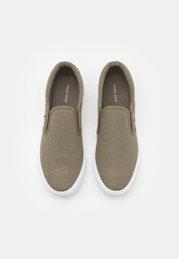 Khaki plátěné slip-on boty s kulatým tvarem, bílé gumové podrážky a hnědými akcenty. Obsahuje logo štítek v červené, bílé a modré barvě.