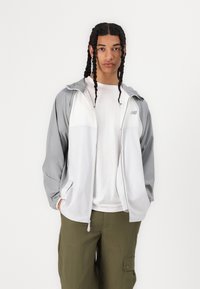 Jachetă zip-up ușoară în gri și alb, cu glugă, două buzunare laterale și un logo New Balance pe piept. Textură netedă.