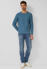 Mann steht vor einer weißen Wand, trägt einen blauen Langarmpullover, ausgewaschene blaue Jeans und weiße Turnschuhe und blickt geradeaus.