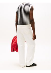 Czarnobiały dzianinowy top bez rękawów w paski, białe spodnie o szerokich nogawkach, trzyma czerwoną pikowaną kurtkę, czarne buty, jednolite tło.