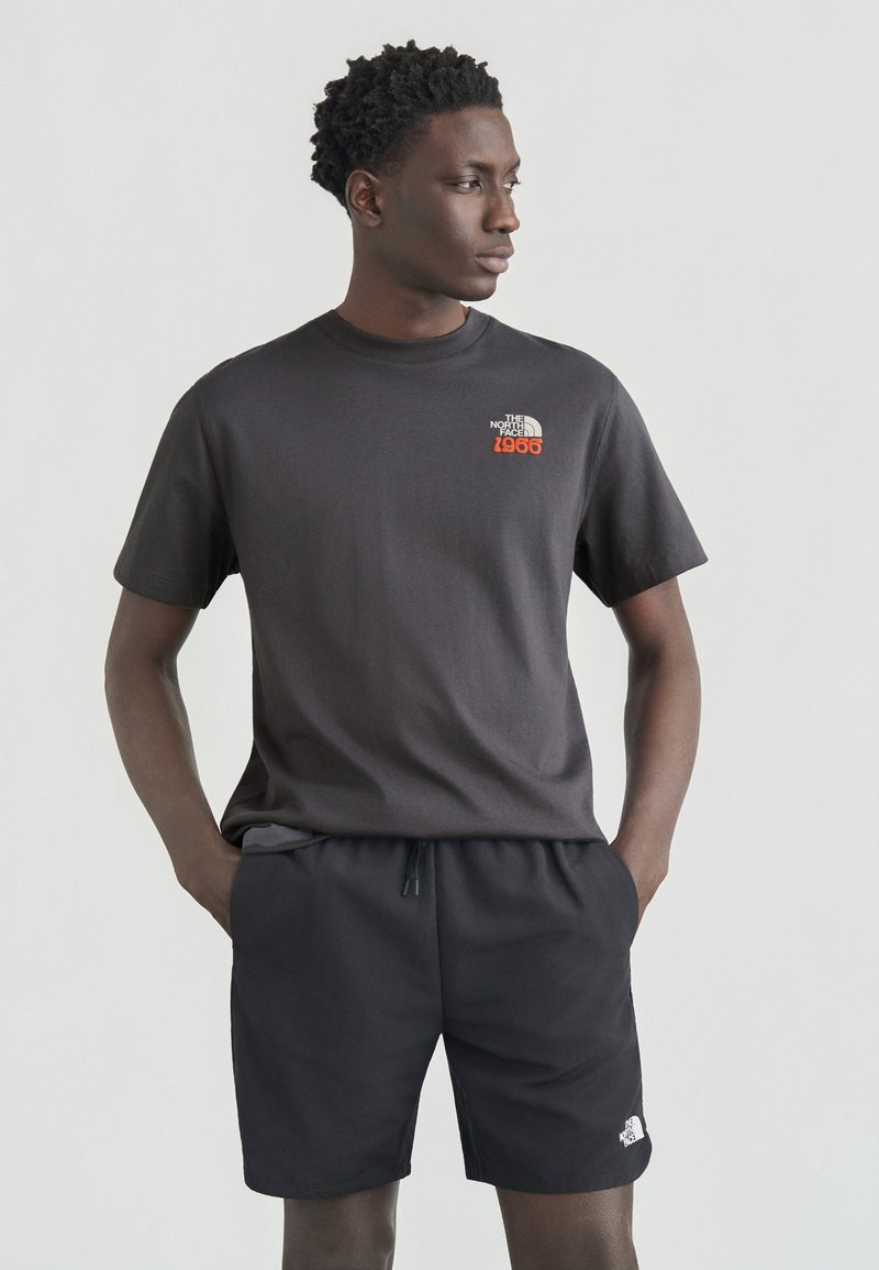 Νέος άνδρας που φοράει μαύρο T-shirt και σορτς The North Face, στέκεται με τα χέρια στις τσέπες, κοιτάζοντας προς τα πλάγια μπροστά σε μια απλή φόντο.