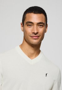 V-Ausschnitt-Pullover aus cremefarbener Maschenware, mit einem kleinen schwarzen Emblem auf der linken Brust. Glatte Textur mit geripptem Halsausschnitt und Bündchen.