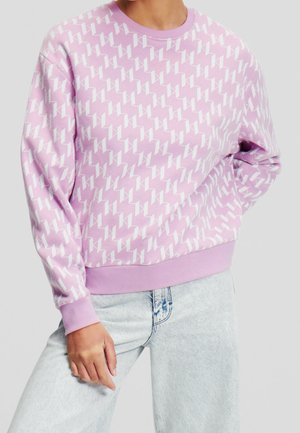 Personne portant un sweat-shirt violet clair avec un motif géométrique blanc et un jean bleu clair, debout avec une main dans la poche.