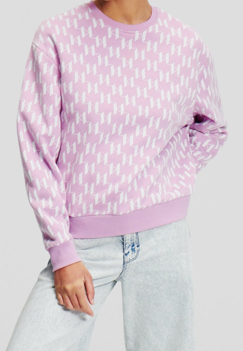 Personne portant un sweat-shirt violet clair avec un motif géométrique blanc et un jean bleu clair, debout avec une main dans la poche.