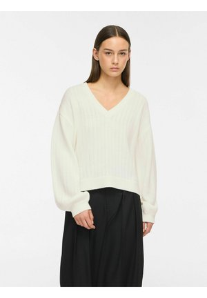 Jeune femme aux cheveux bruns et raides portant un pull blanc à col en V à torsades et un pantalon noir à jambes larges, debout devant un fond blanc.