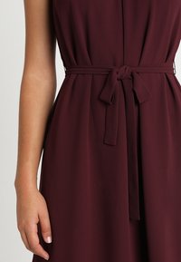 Burgunderfarbenes Kleid mit einem Bindegürtel in der Taille, aus glattem Stoff gefertigt, mit einem gefalteten Design und einem knopfverschlossenen Ausschnitt.