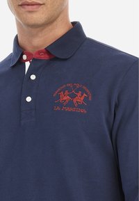 Marineblauw polo shirt van katoen met een rode kraag. Voorzien van witte knopen en een geborduurd logo met daarop twee polo spelers en tekst.