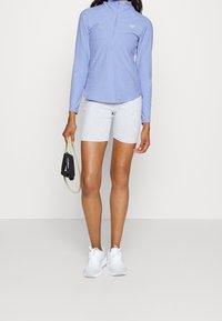 Ljusblå långärmad zip-up-top, grå åtsittande shorts, håller en svart och grön motståndsband, bär vita sneakers med rosa detaljer.