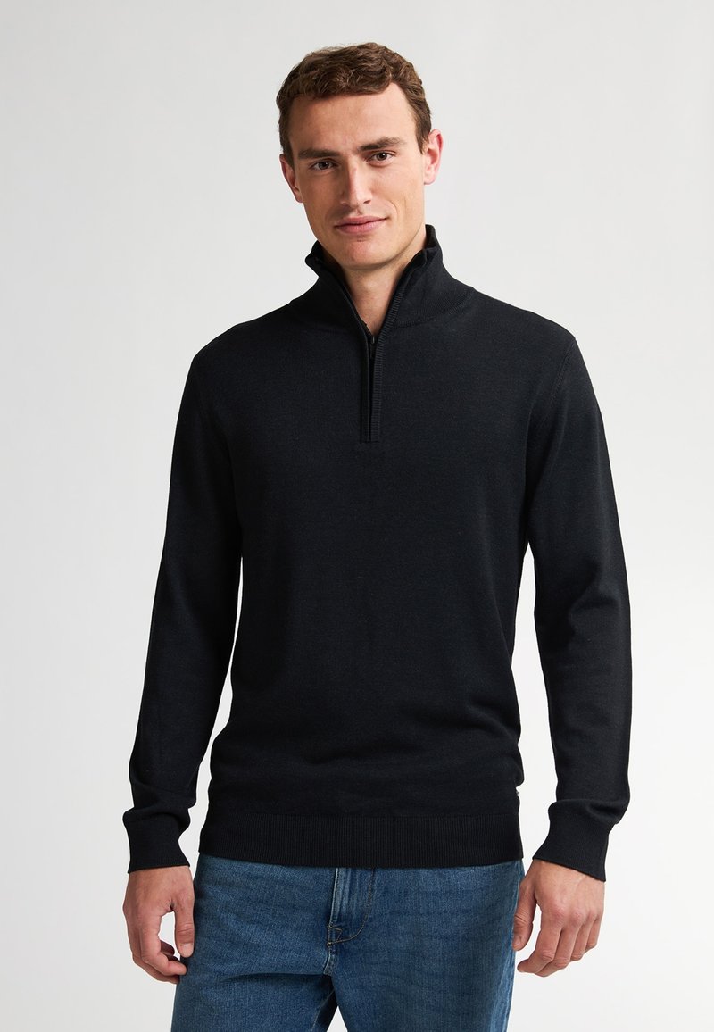 Schwarzer Zip-Kragen-Pullover mit langen Ärmeln, gefertigt aus einem glatten Strickstoff. Verfügt über einen gerippten Kragen und Bündchen, kombiniert mit blauen Denim-Jeans.