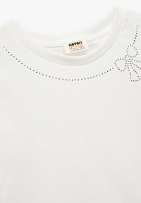 Weißes Baumwoll-T-Shirt mit Rundhalsausschnitt, verziert mit silbernen Strasssteinen in Form eines Schleifenmusters und einer dekorativen Linie entlang des Halsausschnitts.