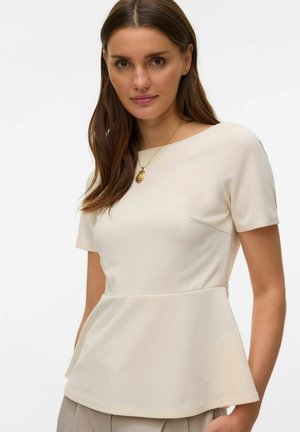 Kvinne med langt brunt hår som har på seg en hvit kortermet peplum-topp og et gullanheng, poserer foran en ensfarget bakgrunn.