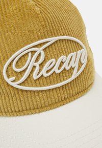 En sennepsgul kordfløjlskasket med hvidt broderet "Recap" logo, der har en struktureret krop og en glat hvid skygge.