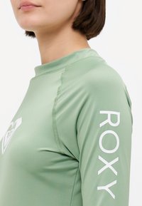 Lys grønne langermede rash guard med rund hals. Har hvit "ROXY"-tekst nedover høyre erme og glatt, elastisk stoff.