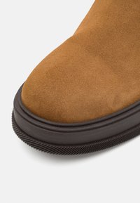 Botte en daim marron avec un bout arrondi, une tige texturée, et une semelle en caoutchouc noire avec des détails rainurés pour l'adhérence.