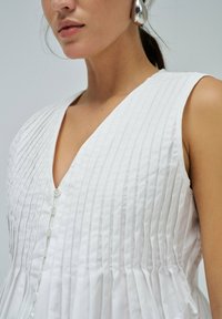 Vestido blanco plisado con escote en V profundo y rayas verticales. Presenta botones en el frente y un dobladillo ligeramente acampanado. Textura suave.