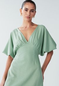 Mintgrön klänning med djup V-ringning och flödande korta ärmar. Har rynkad detaljering vid bysten, tillverkad av mjukt tyg.