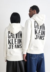 Felpe bianche con cappuccio e grande testo nero "CALVIN KLEIN JEANS" sulla schiena. Una persona indossa una gonna in pelle, l'altra un paio di jeans blu.