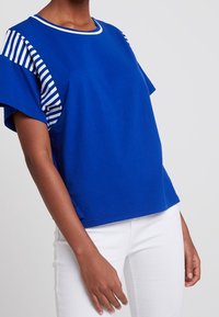 Blue cotton t-shirt with striped sleeves, round neckline, and short sleeves. Soft texture with a relaxed fit. Paired with white pants.