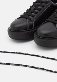 Svarta lädersneakers med svarta platta snörningar och vit söm. Har en strukturerad gummisula och en enkel, rundad design.