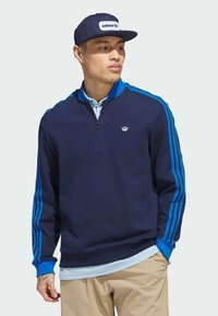 Navy athletic sweatshirt with a quarter zip, blue accents on sleeves, and small logo on chest. Paired with light blue collar and beige pants.