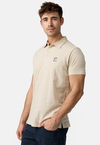 Bež polo majica izrađena od laganog, teksturiranog materijala. Ima klasičan ovratnik i mali crni logo na prsima. U slim-fit dizajnu.