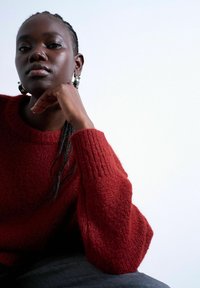 Personne noire aux cheveux tressés portant un pull en tricot rouge, reposant le visage sur la main, devant un fond clair uni.