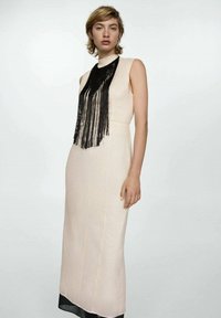 Frau in einem ärmellosen beige Maxi-Kleid mit weißem Kragen und langer schwarzer Quasten-Halskette, die vor einem schlichten weißen Hintergrund steht.