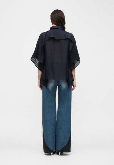 Γυναίκα που στέκεται με την πλάτη προς τα εμπρός, φοράει μια χαλαρή μαύρη μπλούζα με φαρδιά μανίκια και ψηλόμεσα τζιν παντελόνια με φαρδιά πόδια και σκοτεινές καμπυλωτές πινελιές στις άκρες.