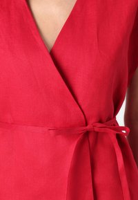 Top avvolgente in lino rosso con scollatura a V profonda e vita annodata. Presenta maniche corte e un tessuto liscio e strutturato in un colore uniforme.