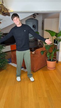 Άτομο που φοράει μαύρο πουλόβερ με ριπ ύφανση και ανοιχτό πράσινο παντελόνι στέκεται σε έναν ζεστό εσωτερικό χώρο με καφέ καναπέ και γλαστράκι.