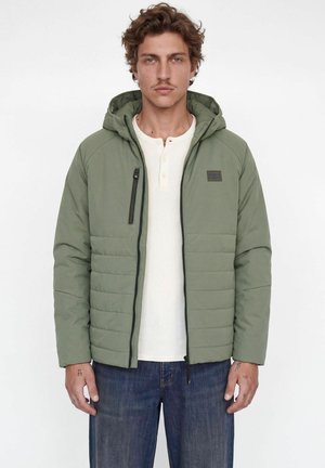 Homme aux cheveux bouclés portant une veste matelassée à capuche verte par-dessus une chemise henley blanche et un jean bleu, debout sur fond blanc.