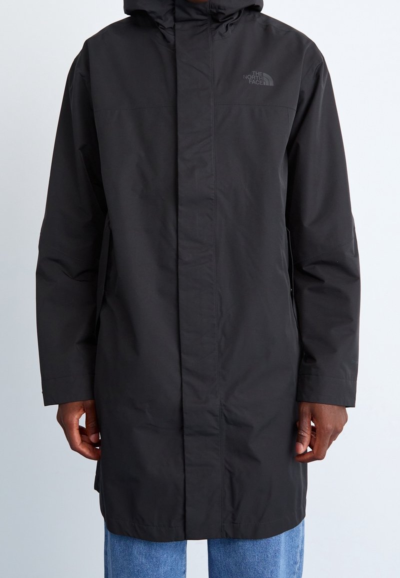 Imperméable long noir The North Face avec capuche, porté par-dessus un jean bleu, vue de face montrant la fermeture éclair fermée et les poches latérales.