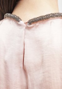 Ljusrosa satängtyg med en detalj av pärlor vid halslinningen. Kragen har olika metallpärlor i ett kontinuerligt mönster.