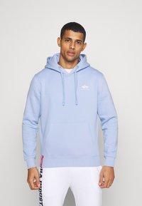Felpa blu chiaro realizzata in tessuto morbido, con tasca frontale, coulisse e un piccolo logo. Abbinata a pantaloni bianchi con logo.