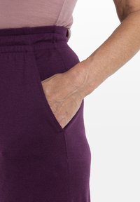 Lila, avslappnad passform, byxor med elastisk midja och sidficka i mjukt, strukturerat tyg. Närbild visar en hand som vilar i fickan.