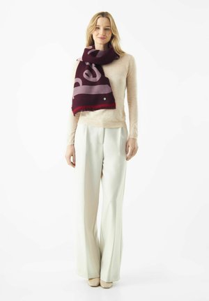 Rode en bordeauxrode sjaal met grote letters, gedragen over een lichte trui en witte wijde broek. Soepele textuur met franjes aan de randen.