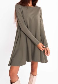 Femme portant une robe ample à manches longues de couleur vert olive, associée à des bottes beiges claires jusqu'aux genoux, debout devant un fond blanc uni.