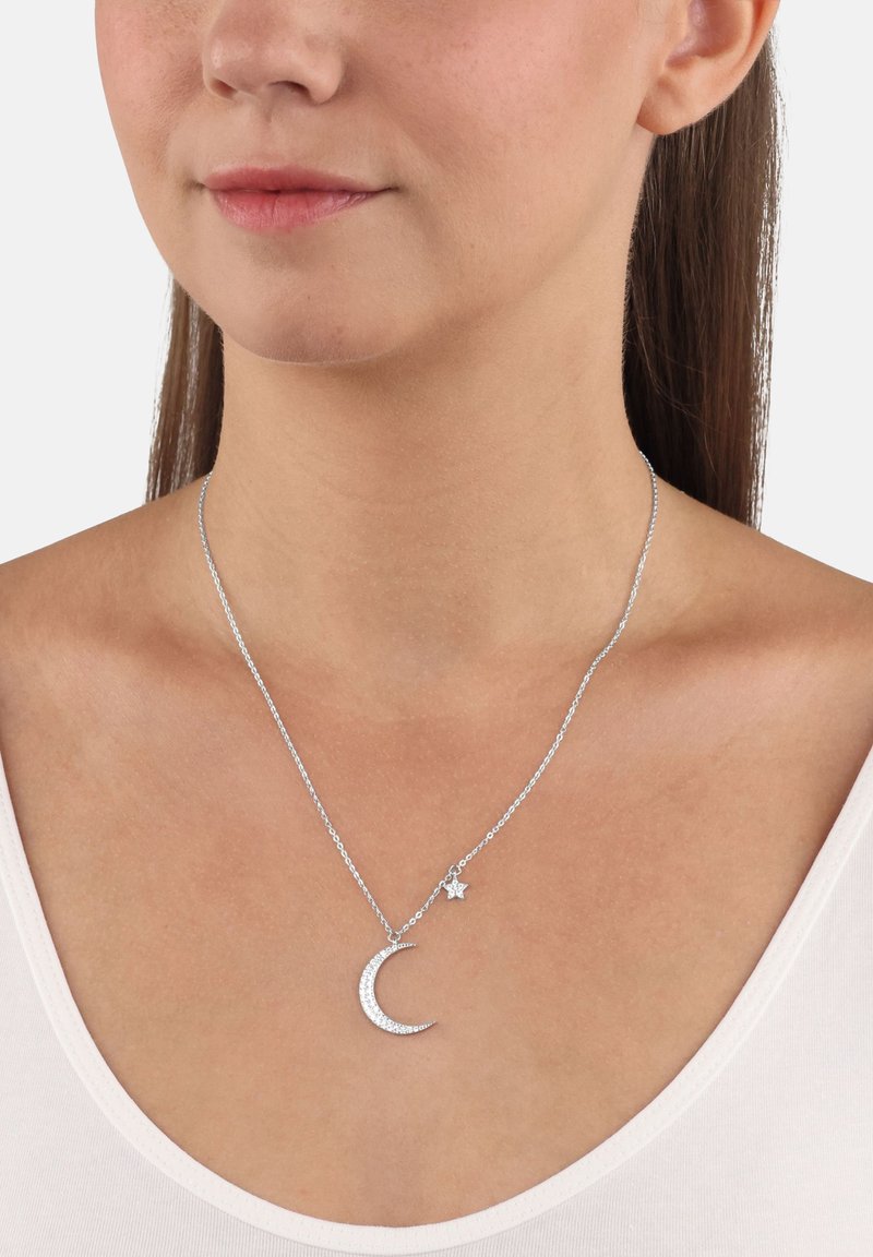 Collana d'argento con pendente a forma di luna crescente e stella, adornata con pietre trasparenti, attaccata a una catena sottile, indossata sopra una maglietta chiara.